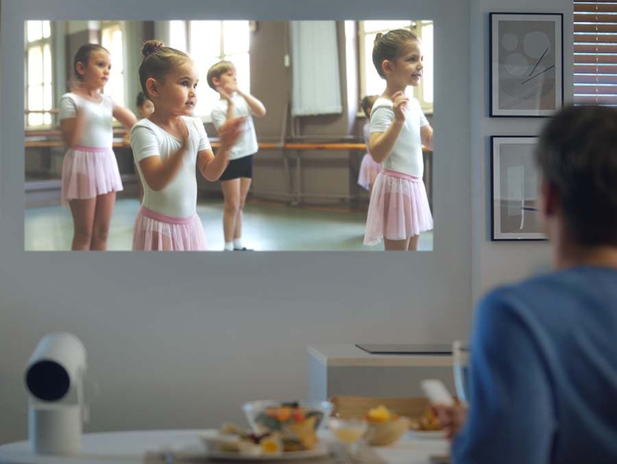 A man is playing a video of children in a ballet class with The Freestyle. The video is slightly obstructed by frames hanging on the wall, but The Freestyle detects this and automatically moves the projection away from the frames.