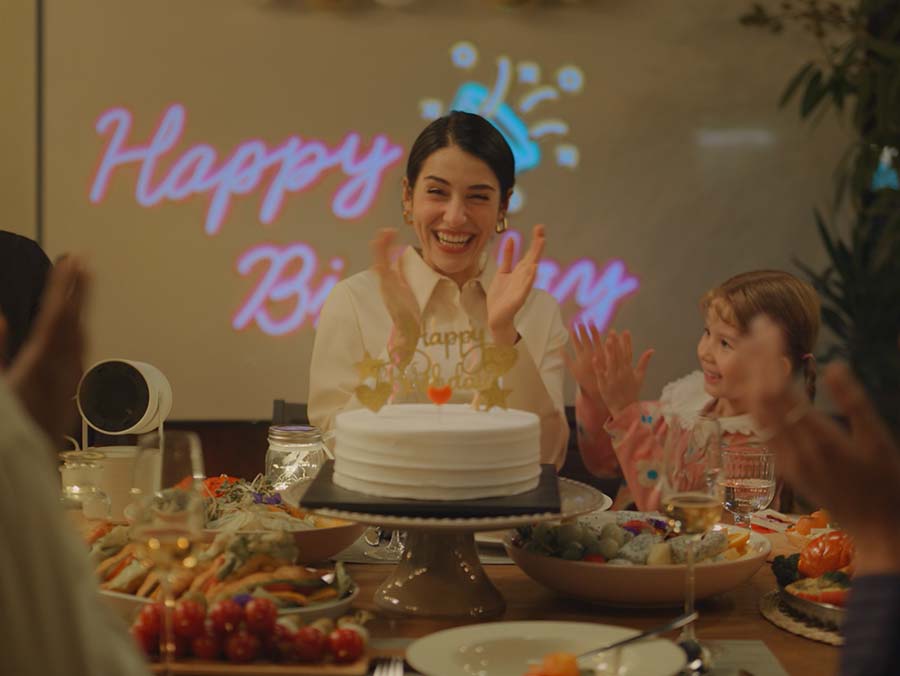 A happy woman surrounded by people blows out candles on a cake. Behind her is a happy birthday decoration projected by The Freestyle on the wall. The camera zooms out, revealing that the party is outdoors on a terrace next to a swimming pool.