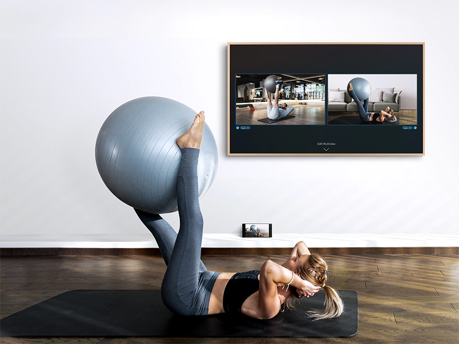 A woman is working out with an exercise ball on the ground with her smartphone next to her. The Frame is displaying what's on her smartphone as well as a video of a trainer on the TV's screen. LS03BBUXXH