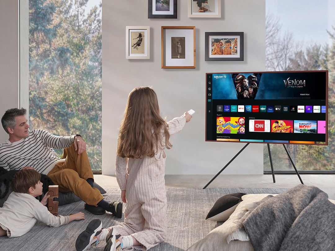 A girl is pointing the remote towards The Frame, which shows the Smart Hub home screen. LS03BBUXXH